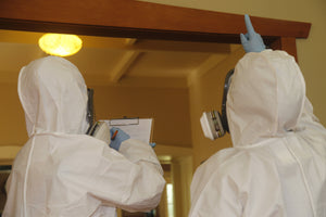 Person in white hazmat suit taking asbestos notes on a clipboard while another person in a white hazmat suit points at a building material during an asbestos survey in a house