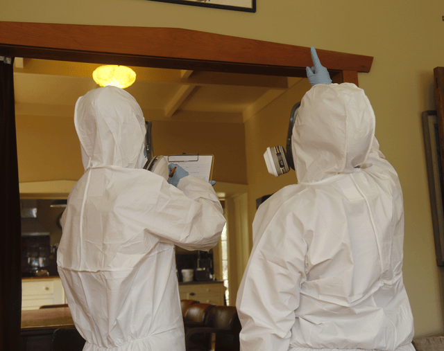 Person in white hazmat suit taking asbestos notes on a clipboard while another person in a white hazmat suit points at a building material during an asbestos survey