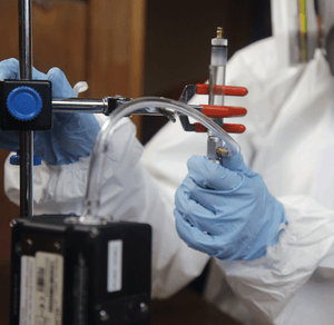 Person in white hazmat suit, blue gloves, and respirator holding sampling tube connected to a pump to collect indoor air quality samples