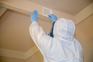 Person in white hazmat suit and blue gloves collecting a swab sample from a ceiling in a residential home during a meth test